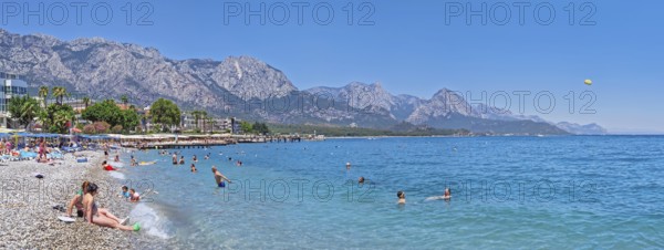 KEMER, TURKEY - JULY 5, 2025 Kemer municipality public beach with vibrant pebble under a clear, turquoise sea water. People swimming and sunbathing on the shore. Beautiful Taurus mountains at horizon