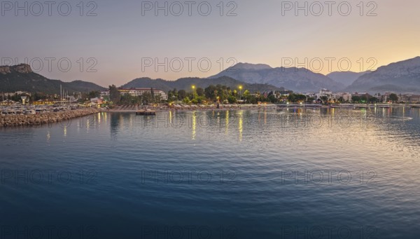 Dusk scene in Kemer, Turkey, showing the town illuminated coastline and beach, reflected beautifully on the calm sea. Majestic Taurus mountains silhouettes in the background