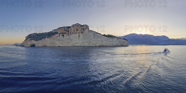 Vibrant evening seascape with a boat cruising away on the blue sea, near the Pirate Cave in Kemer at sunset. Beautiful Turkish mediterranean coastline lined with tree covered rocky cliffs at dusk