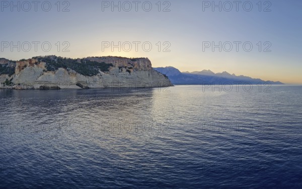 Vibrant seascape sunset with a view to the Pirate Cave in Kemer, Turkey. Beautiful Turkish mediterranean coastline lined with tree covered rocky cliffs and hills under a clear evening sky