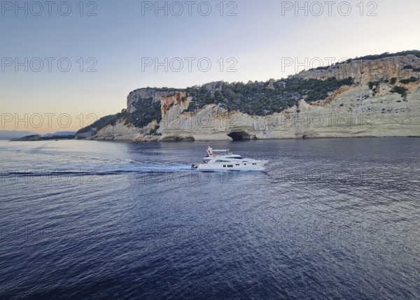 Vibrant evening seascape with a boat cruising away on the blue sea, near the Pirate Cave in Kemer at sunset. Beautiful Turkish mediterranean coastline lined with tree covered rocky cliffs at dusk