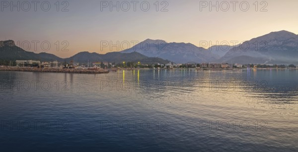 Dusk scene in Kemer, Turkey, showing the town illuminated coastline and beach, reflected beautifully on the calm sea. Majestic Taurus mountains silhouettes in the background