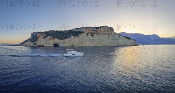 Evening seascape panorama with a boat cruising away on the blue sea, near the Pirate Cave in Kemer at sunset. Beautiful Turkish mediterranean coastline lined with tree covered rocky cliffs at dusk