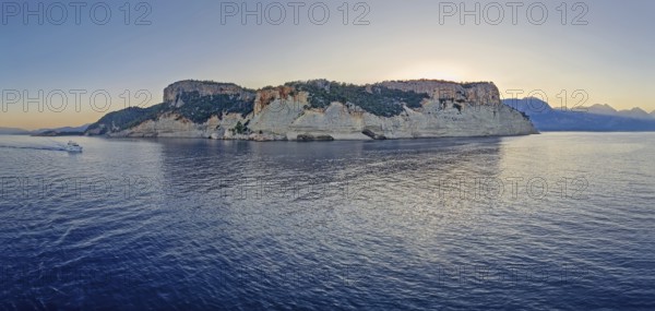Evening seascape panorama with a boat cruising away on the blue sea, near the Pirate Cave in Kemer at sunset. Beautiful Turkish mediterranean coastline lined with tree covered rocky cliffs at dusk