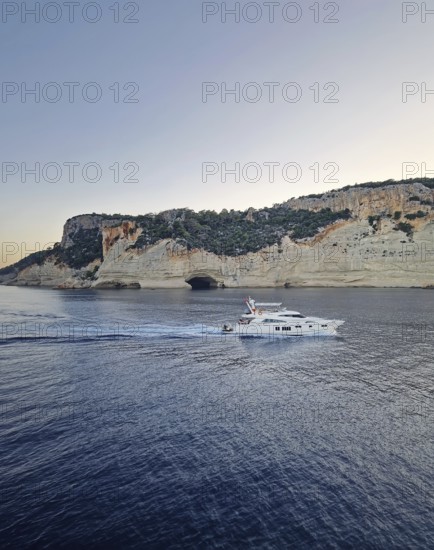 Vibrant evening seascape with a boat cruising away on the blue sea, near the Pirate Cave in Kemer at sunset. Beautiful Turkish mediterranean coastline lined with tree covered rocky cliffs at dusk