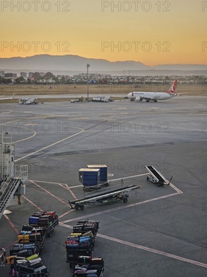 ANTALYA, TURKEY - JULY 5, 2025 Antalya airport scene at sunset with luggage being moved onto carts in the foreground and a plane on the tarmac. Distant mountains range silhouette in the background