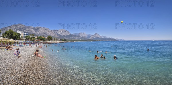 KEMER, TURKEY - JULY 5, 2025 Kemer municipality public beach with vibrant pebble under a clear, turquoise sea water. People swimming and sunbathing on the shore. Beautiful Taurus mountains at horizon