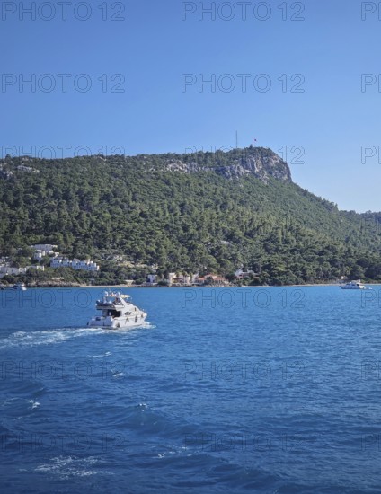Vibrant seascape with a boat cruising away on the blue sea, leaving a white wake. Beautiful Turkish mediterranean coastline lined with tree covered Calis hill under a clear sky in Kemer, Turkey