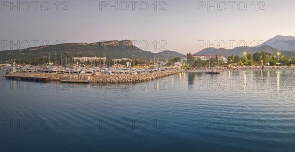 Kemer marina at dusk, filled with multiple docked boats. Lights from the harbor and hotels on the resort shoreline reflects on the calm sea water