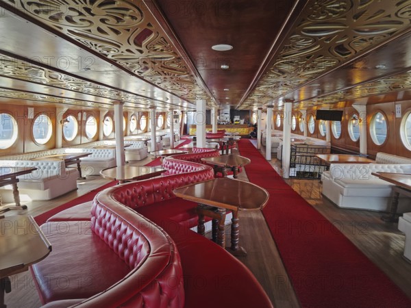 Luxurious ship interior with long hall, red carpets, leather couches and curved wooden tables. Ornate golden patterns adorn the ceiling, and circular windows line the walls. Vintage charm and comfort