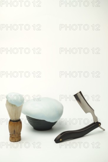 Shaving foam in a bowl, shaving brush and a straight razor are arranged on a white background, composing a still life focused on men's grooming