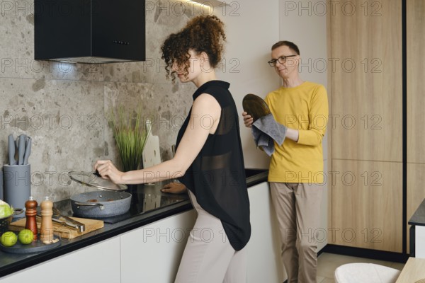 A woman stirs a pot on the stove while a a man wipes the washed dishes and watches her in a stylish kitchen
