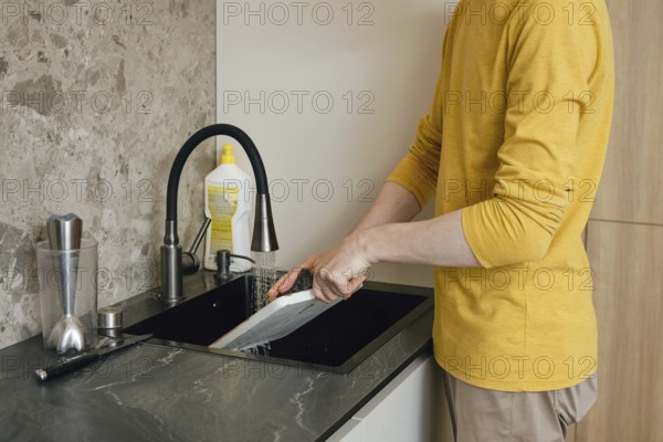 A person is cleaning a chopping board under running water in a sleek modern kitchen. The individual is dressed in a yellow long-sleeve shirt, focused on washing the dishes thoroughly