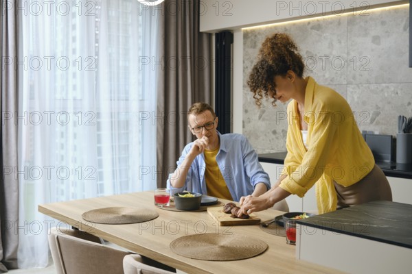 A man and woman are preparing a meal together in a modern kitchen. The woman slices food on a wooden cutting board while the man observes, smiling