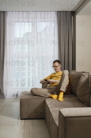 A man sits comfortably on a stylish couch in a contemporary apartment. He is focused on his smartphone, wearing casual attire and bright yellow socks, surrounded by soft drapery and natural light