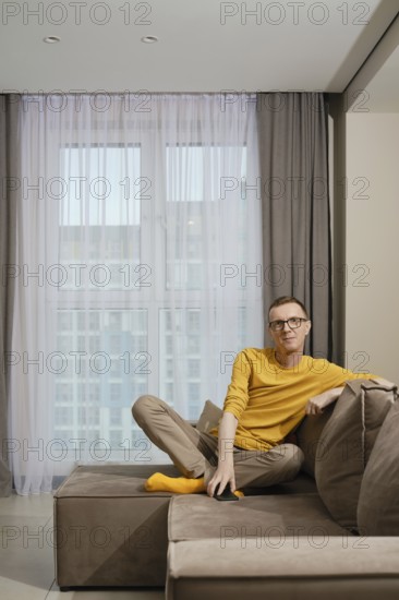 A man wearing a yellow shirt and socks and beige pants sits comfortably on a brown couch, relaxed in a well-lit modern apartment with large windows and curtains
