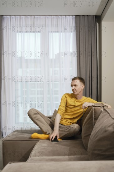 A dreamy man dressed in a yellow shirt and beige pants sits comfortably on a sofa, casually resting his leg in yellow socks