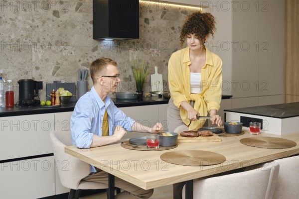 In a contemporary kitchen, a couple is sharing a moment as one partner serves a meal while the other is seated. The setting features stylish decor and a warm atmosphere, perfect for dining together