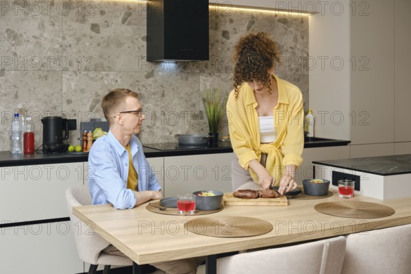 A woman slices roasted duck breast on a cutting board while a man sits at the dining table. They share a relaxed afternoon together, enjoying a homemade meal and drinks