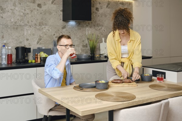 A man sits at a wooden dining table, sipping a drink, while a woman prepares a meal on the countertop in a stylish kitchen. The atmosphere is cozy and lively, emphasizing togetherness