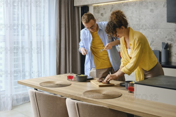 In a bright kitchen, a couple is joyfully preparing a meal together. The woman slices meat on a wooden board. The modern space is filled with natural light