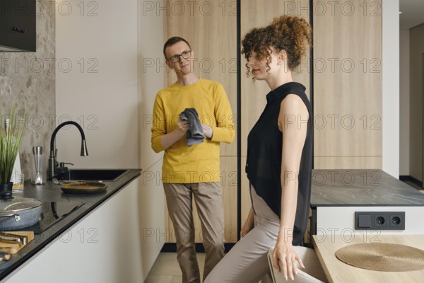 In a stylish kitchen, a man in a yellow shirt looks thoughtfully at a woman in a black top as she poses leaning on table. Their relaxed atmosphere suggests a shared cooking experience