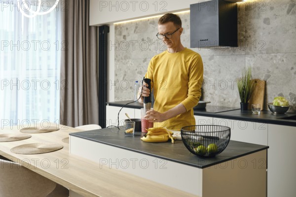 A middle-aged man prepares banana and strawberry smoothies in the kitchen using an immersion blender