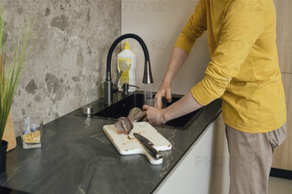 A person wearing a yellow shirt is washing utensils and a cutting board in a dark kitchen sink. The modern kitchen has a stone backsplash, while the individual carefully uses dish soap for cleaning