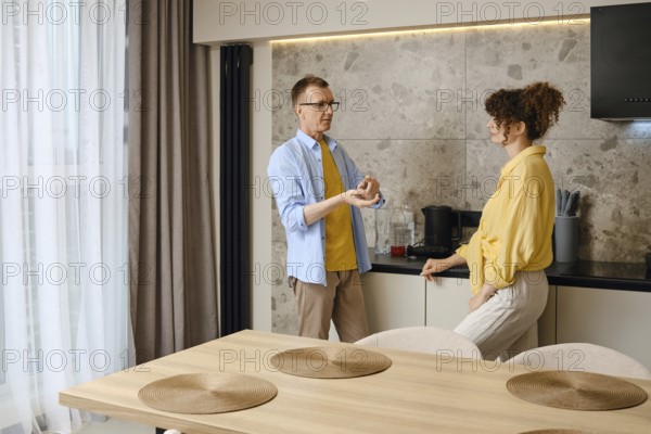 A couple engages in conversation near a kitchen counter, discussing ideas as they prepare for a meal. The modern kitchen is bright and airy, enhancing the cozy atmosphere of their interaction