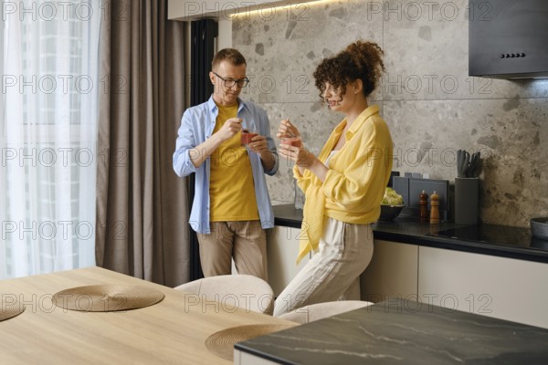 A couple stands in a stylish kitchen and eating strawberry and banana smoothies. Sunlight filters through the window, enhancing the warm atmosphere of their intimate moment