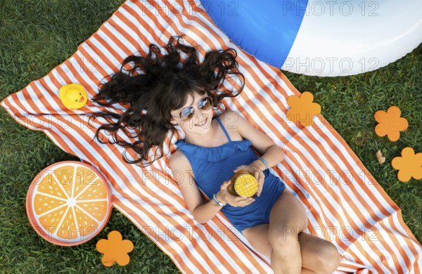 Girl, 10, with sunglasses in a blue swimming costume, lying relaxed in the meadow with a cocktail on a towel