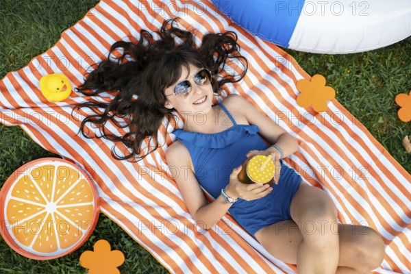Girl, 10, with sunglasses in a blue swimming costume, lying relaxed in the meadow with a cocktail on a towel