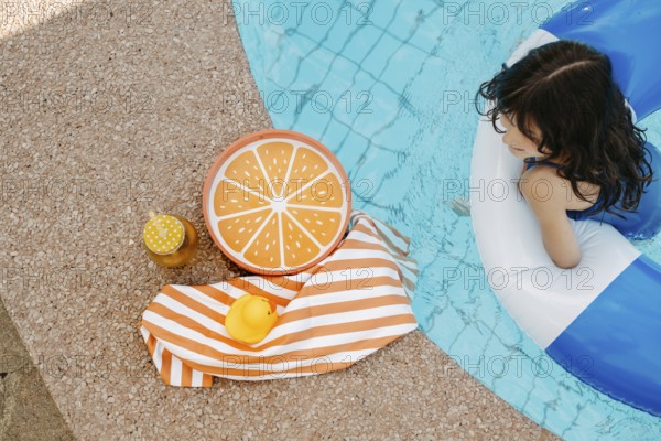 Girl with a swimming tyre in the pool, a striped bath towel, a swimming duck, a cocktail and an orange tray at the edge of the pool