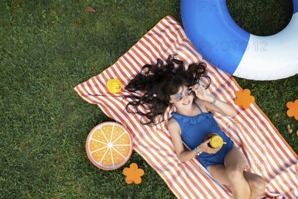 Girl, 10, with sunglasses in a blue swimming costume, lying relaxed in the meadow with a cocktail on a towel