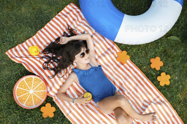 Girl, 10, with sunglasses in a blue swimming costume, lying relaxed in the meadow with a cocktail on a towel