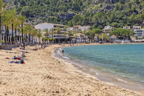 Sand beach with tourists at a tourist resort with houses at the shore line and palm trees a sunny summer day, Port de Sóller, Mallorca, Spain