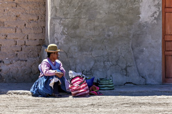 Woman in traditional dress sitting on the floor next to colourful bags in front of a wall, Traditional woman in a village in the Salar de Uyuni in Bolivia
