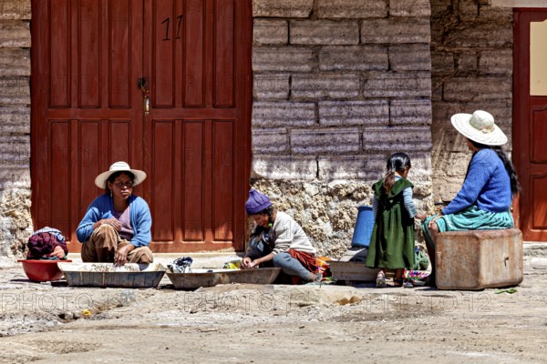 Women and children sit in front of a stone house, busy with housework. The sun shines brightly, traditionally dressed woman and children sit on the ground in the Salar de Uyuni in Bolivia