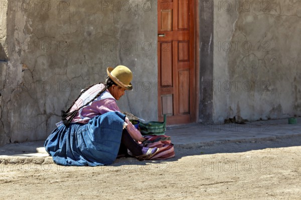 A woman in traditional dress sits on the street in front of a wall in the sun, Traditionally dressed woman sits on the ground in the Salar de Uyuni in Bolivia