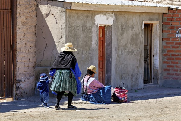 Woman and child in traditional dress standing in front of a brick house on a sunlit street, Traditional woman in a village in the Salar de Uyuni in Bolivia