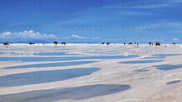 Puddles on a salt flat with vehicles on the horizon and clouds in the sky, The endless expanses of the salt plain of the Salar de Uyuni in Bolivia