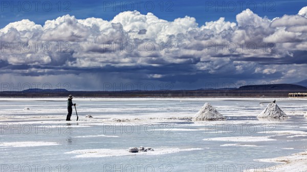A single figure shovels salt under a dramatic cloudy sky on vast salt flats, The endless expanses of the salt plain of the Salar de Uyuni in Bolivia