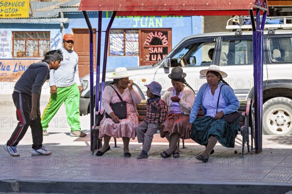 A group of people in traditional dress sit on a bench surrounded by shops and a car, people at a bus stop in Uyuni, Bolivia