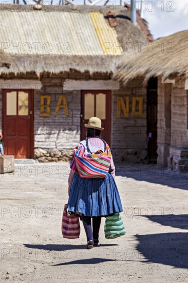 A woman in colourful traditional dress carries bags in a village with rustic buildings, Traditional woman in a village in the Salar de Uyuni in Bolivia