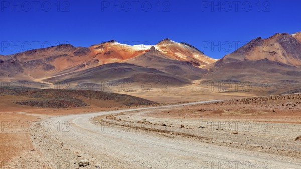 A winding sandy road through a dry landscape with reddish mountain peaks and blue skies, The landscape of the Altiplano in Bolivia