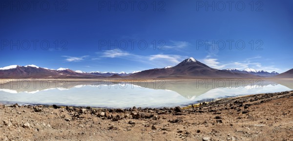 Wide view of a volcano with a reflecting lake and rocky shore under a blue sky, The Laguna Colorada in Bolivia
