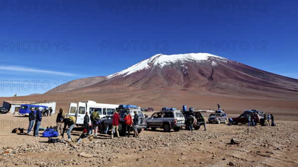 Groups of people and vehicles in a desert landscape with a snow-covered mountain under a blue sky, Off Road in the Landscape of the Altiplano in Bolivia