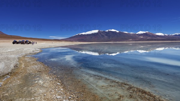 Panoramic view of a lake with snow-capped peaks, people and vehicles on the shore, The Laguna Colorada in Bolivia