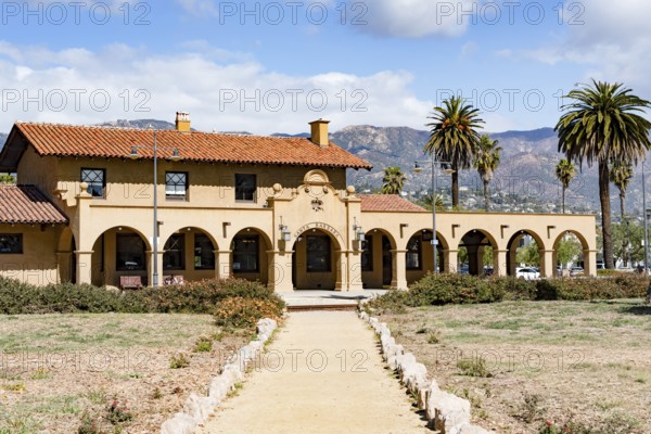 The historic Santa Barbara train station, featuring Spanish Colonial Revival architecture with a red tile roof and arches, set against scenic mountains and palm trees, California, USA