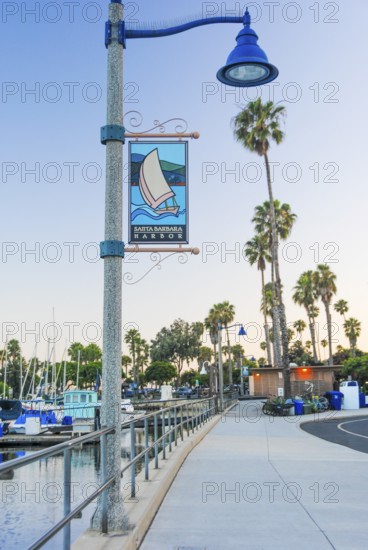 A serene view of Santa Barbara Harbor at dusk, featuring a decorative street lamp with a banner, a walkway, and sailboats in the background, California, USA